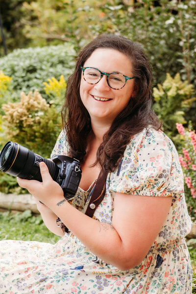 photo of philadelphia maternity photographer, Kristi, in front of a blooming garden bed.  Kristi is sitting and smiling while holding her nikon d750 camera and 24-70mm lens