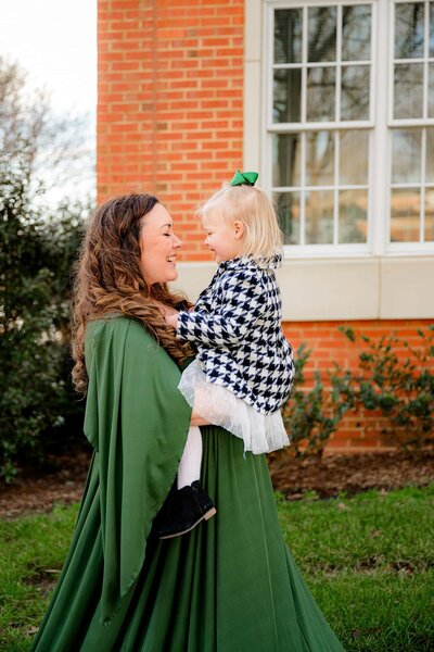 An expectant Charlotte mom standing on the University of North Carolina Charlotte's campus holding her daughter mother is wearing a dark green maternity dress and her blonde daughter is wearing a houndstooth coat