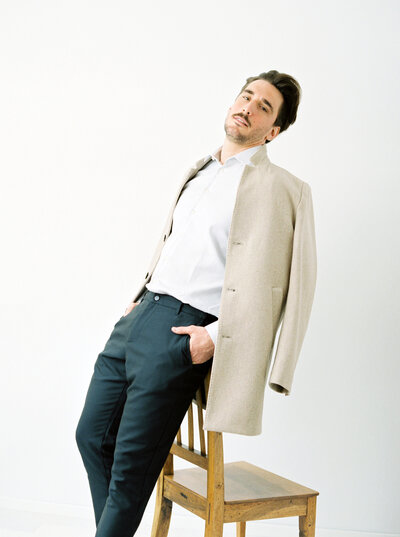 Man poses for portrait leaning against a chair in cream overcoat