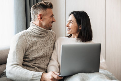 A Couple smile at one another as they listen to something on their laptop. This could symbolize what a teletherapy session might look like from the comfort of your home. We offer online marriage counseling, online relationship therapy, and more.