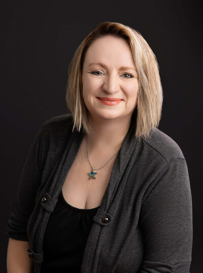 A woman with shoulder-length blond hair smiles at the camera. She is wearing a dark top with a cardigan and a necklace with a star-shaped pendant. The background is dark, accentuating her bright attire and friendly expression, much like the warmth you'll find in any Jacksonville newborn photographer's portfolio.