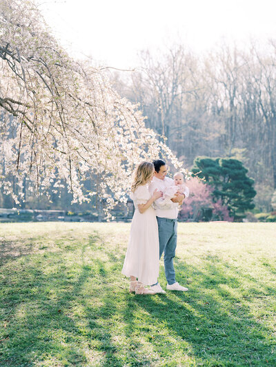 Mother and father stand holding baby girl in front of cherry blossom tree at Brookside Gardens.