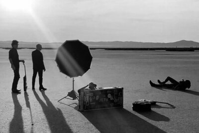 black and white bts photo Mark Maryanovich lying on ground while photographing 7Horse standing in desert guitar and case beside reflector in front of them