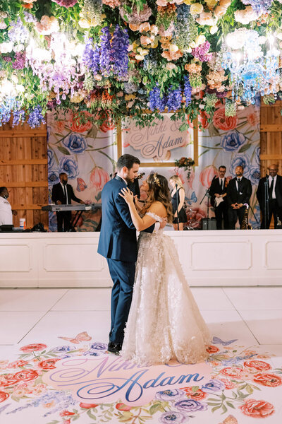 Bride and groom first dance under colored flowers and pastel chandeliers.