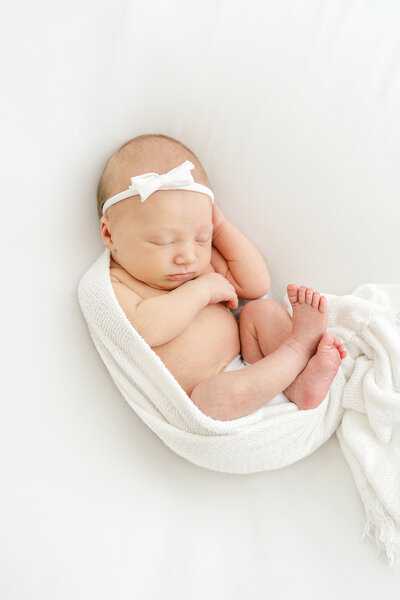 Newborn baby girl posed on white blanket and wearing tiny white hair bow during newborn photo shoot at Julie Brock Photography in Louisville KY
