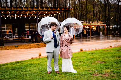 Bride and groom standing under clear umbrellas in front of the Meadows Raleigh wedding venue