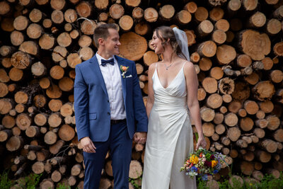 A bride and groom stand in front of a large pile of trees holding hands and looking at each other while she holds her bouquet in the other hand.