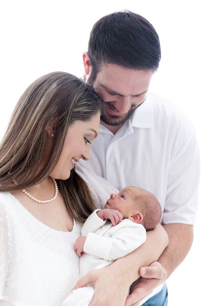Happy parents smile down at their awake newborn baby while standing in a studio in all white clothes