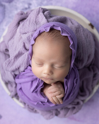Sleeping newborn girl wearing purple  wrap and bonnet with ruffles, on purple background, with hands together under chin