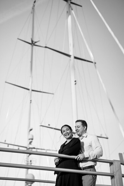 A couple dressed in elegant attire, the woman in black and the man in a white shirt, leaning against a railing with a large yacht in the background