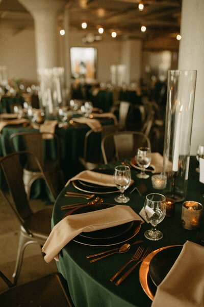 Elegant banquet setting with dark green tablecloths, gold cutlery, and tall glass vases, in a warmly lit hall, meticulously arranged by an Iowa wedding planner.
