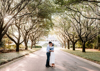 man and woman kissing under large trees while standing in long driveway