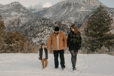 A family of three walks together in the snow with mountains and pine trees in the background, capturing a heartwarming Colorado family lifestyle session in the wild.
