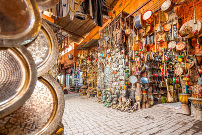 A vibrant market stall displaying a variety of colorful musical instruments, including drums, tambourines, and stringed instruments. The setting is open-air with a cobblestone floor and rustic orange walls.