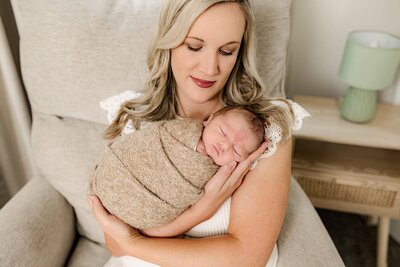 Mother snuggles her newborn son and her arms while sitting in a chair during their lifestyle newborn session