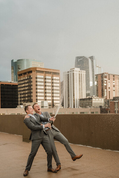 Couple pops a bottle of champagne after their engagement with views of downtown Denver in the background