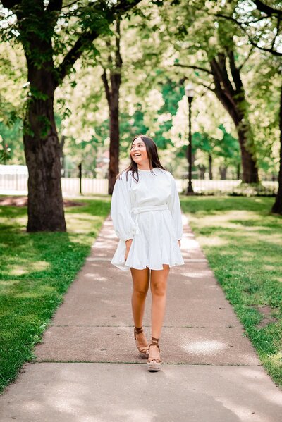 Senior girl wearing white dress and walking down the sidewalk laughing