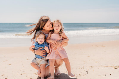 Mom kneeling down and holding her two young children on the beach.  Photo taken by Bethany Beach family photographer, Kristi