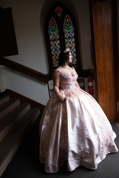 A teenage girl in a tiara and pink ballgown stands in front of a stained glass window Rockland County quinceanera photographer Milestone Images