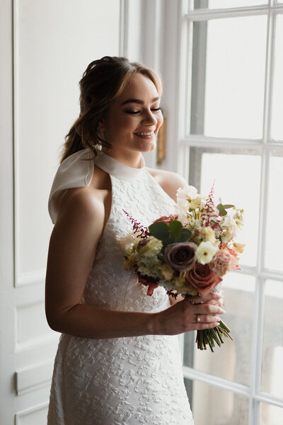 Bride stands near window holding her bouquet and laughs with her eyes closed