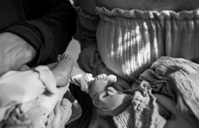 Black and white image of a pregnant mother and her husband holding their newborn twins. The parents share a heartfelt moment, looking at their baby's tiny feet.