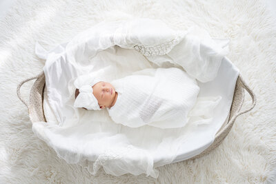 A baby girl in a large headband with a bow, wrapped in a blanket and sleeping in a Moses basket