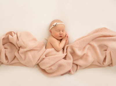 A newborn baby sleeping wrapped in a peach blanket with  a rosette headband
