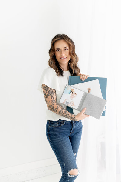 Greater Atlanta Motherhood & Family Photographer Caya holds some fabric covered newborn photo albums while standing in jeans