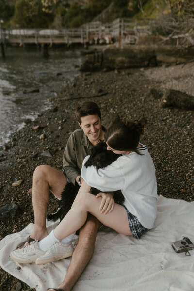 Couple and dog cuddling on white picnic blanket on rocky beach shore with film camera