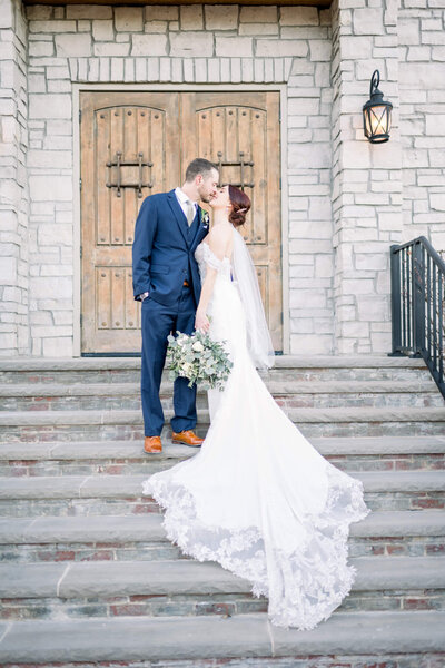man and woman are dressed in wedding attire, kiss in front of wooden door and rock wedding venue
