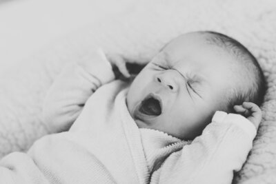 net geboren baby in geboortepakje van prenatal met mutsje op en slofjes aan. De baby ligt op bed en gaapt.