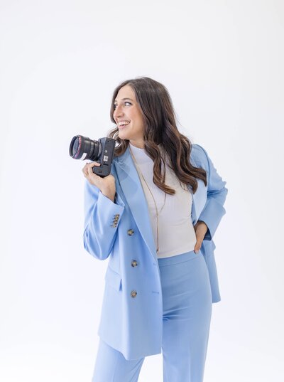 Nashville Wedding Photographer Brooke Elliott posing in a powder blue pant suit and holding her camera close to her face while she looks and laughts out a window.
