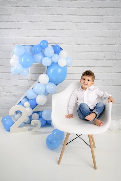 little boy in a white shirt sits in a chair next to a large number 2 filled with balloons