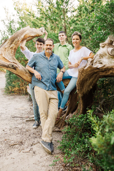 A family of four poses outdoors in front of large tree branches, captured by a Fayetteville NC family photographer. The father stands in front, the mother is on the right, and two sons are in the middle and left. Surrounded by greenery, they appear relaxed and at ease.