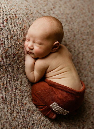 A newborn boy is sleeping while wearing a pair of rust colored pants.