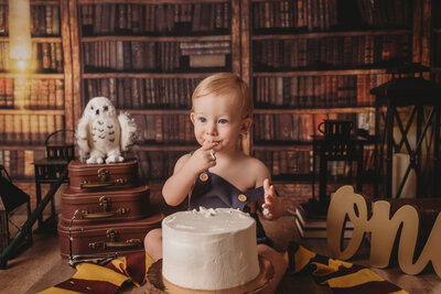 Harry Potter cake smash with one year old little boy eating a cake with Harry Potter owl and bookcase behind him