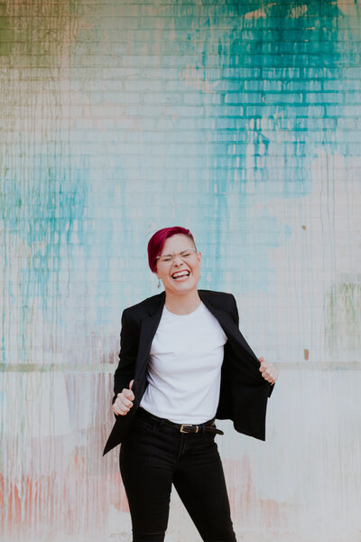Personality headshot of web designer Kassidi Gniadek wearing a black suit laughing in front of colorful background at Camp North End in Charlotte