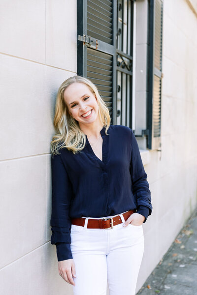 Digital Grace Design owner, Sarah Blodgett, leans against wall outside and smiles wearing a blue silk blouse and white pants