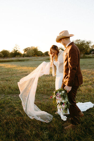 Bride and groom walking off towards sunset with veil flowing and groom in cowboy hat