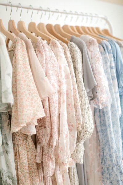 A selection of neutral colored dresses hang on display in the closet of New Jersey Newborn Photographer Kate Voda Photography. The first dress is cream with small light gray flowers, the second is gray with short flutter sleeves, and the third is white with long sleeves.