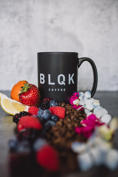 Product photo of black coffee mug surrounded by fruit and flowers