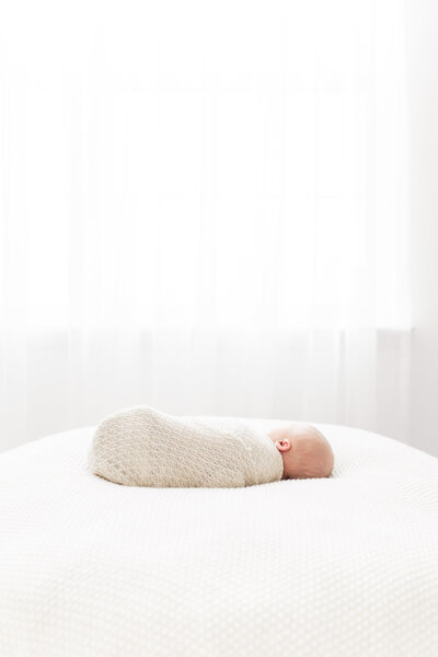 A DC Newborn Photography photo of a newborn baby sitting on a white beanbag in front of white sheer curtains in front of a window