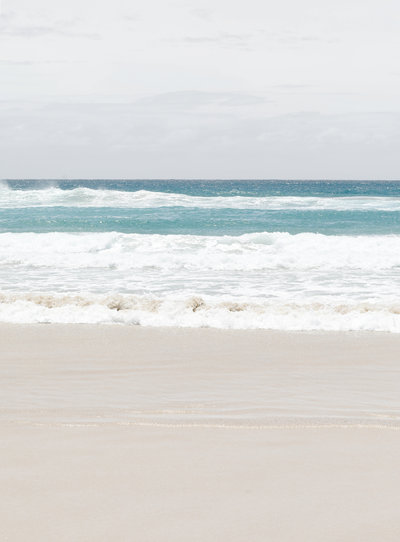 Pristine beach in Byron Bay, Australia. The water is teal and white and the sand is fine and soft. The waves are crawling onto the shore and it's a very peaceful scene.