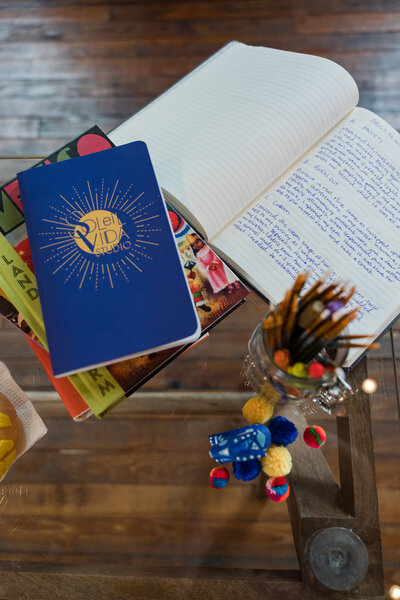 The crafted wonders on our wooden table, showcasing the founder's artistic touch. From books adorned with colorful covers and a blue agenda to the brand's submark and a playful key ring featuring vibrant pompoms, witness the essence of Soleil Vida Studio's creativity.