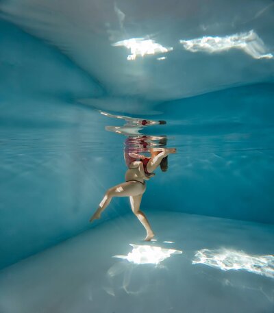 Pregnant woman floating gracefully underwater in a maternity photoshoot, her body reflected on the surface as light dances through the pool.
