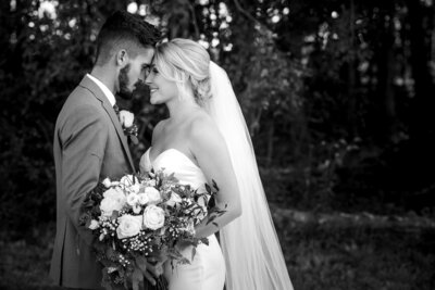 Black and white image of a bride and groom smiling with their foreheads together holding the brides bouquet near the woods at Dove Meadows Estate by Charlotte wedding photographers DeLong Photography