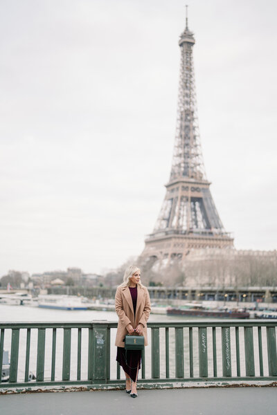 a blonde woman in a tan trench coat holding a green purse looking off into the distance with the eiffel tower behind her