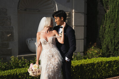 Wedding couple at The Greystone Mansion in California. Bride has a beautiful lace gown while groom has  an elegant tuxedo.  Bride is holding a beautiful blush lush bridal bouquet in her hand.