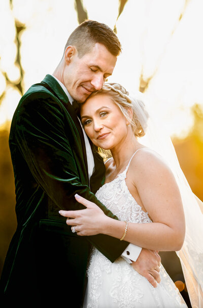 Bride in a white gown and a groom in a green velvet tux jacket standing outside of Hamilton Manor at sunset in the Fall