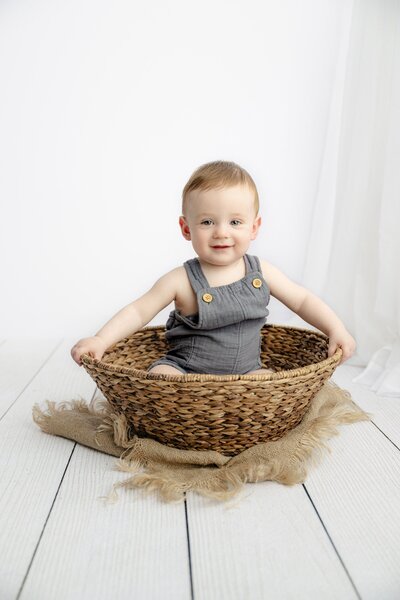 7 month old sits in a basket wearing a blue romper during his waxhaw portrait studio session